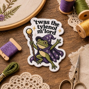 May include: An embroidered patch featuring a green frog dressed as a wizard in a purple star-patterned cloak and hat, holding a staff with a yellow orb. The text 'twas the tylenol m'lord' is stitched in black above the frog on a white background.