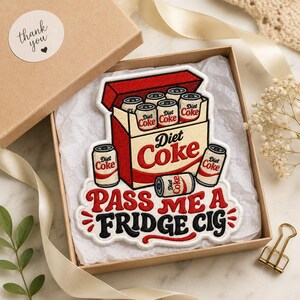 May include: An embroidered patch featuring a red and white Diet Coke carton filled with soda cans, with the text "PASS ME A FRIDGE CIG" in red and black lettering below. The patch is displayed inside a cardboard gift box with white tissue paper.