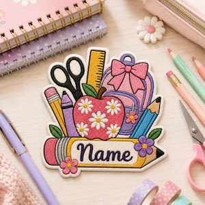 May include: An embroidered patch featuring a red apple with white daisy flowers, a yellow ruler, black scissors, a pink backpack with a bow, and pencils. A horizontal yellow pencil at the bottom includes the word Name in dark purple script text.