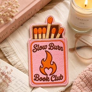 May include: An embroidered patch shaped like a matchbox with a pink background, featuring the text Slow Burn Book Club in brown letters and an orange flame icon. The top section shows six matches with orange tips arranged in a brown box frame.