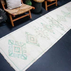 Könnte beinhalten: Ein langer, rechteckiger Läufer mit cremefarbener Basis und sich wiederholenden geometrischen Rautenmustern in sanften mintgrünen und gedeckten beigen Tönen. Der Teppich hat eine strukturierte, gewebte Optik mit Fransen an den Enden.