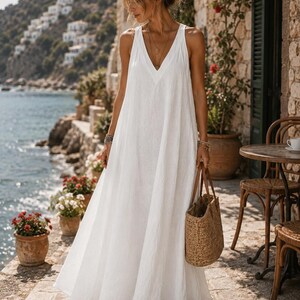 Pode incluir: Um vestido branco comprido sem mangas com um decote em V profundo e uma silhueta larga e fluida. O vestido é feito de um tecido leve e é combinado com uma mala de palha entrançada com pegas em pele castanha.