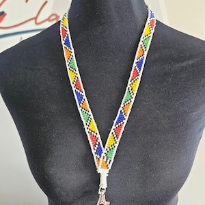 Könnte beinhalten: Ein Schlüsselband aus kleinen Perlen, die in einem geometrischen Muster aus weißen, roten, blauen, grünen und orangefarbenen Dreiecken angeordnet sind. Das Perlenband verfügt über einen silberfarbenen Karabinerhaken aus Metall am unteren Ende.