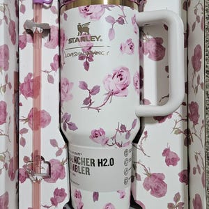 May include: White Stanley Quencher H2.0 tumbler with a pink rose floral pattern, a matching straw, and a bow-shaped straw topper. The tumbler has a handle and is packaged in a floral-printed box.