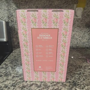 May include: A pink floral patterned box for a Quencher H2.0 Tumbler. The box has a pink background with a repeating pattern of pink roses and green vines. The front of the box has a rectangular label with the product name and features.