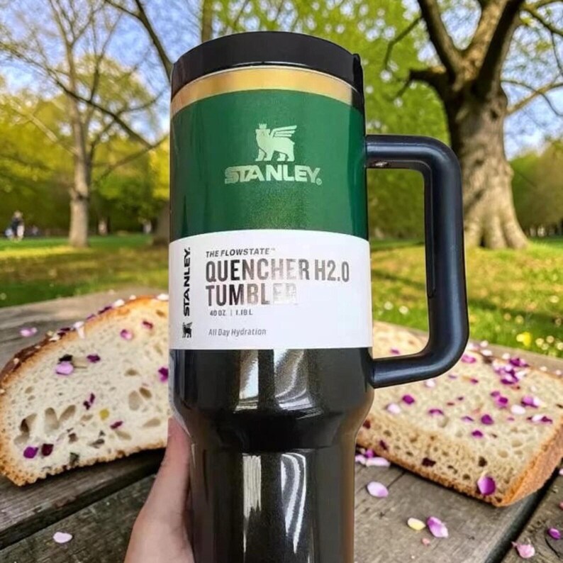 May include: A green and black Stanley Quencher H2.0 tumbler with a black handle. The tumbler has the text "QUENCHER H2.0 TUMBLER" and "All Day Hydration" printed on it. The tumbler holds 40 oz.