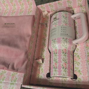 May include: A floral-patterned Stanley tumbler with a handle, straw, and lid, presented in a matching box. The tumbler features a pink and white rose design. The box and accessories are also decorated with the same floral pattern. The tumbler has a capacity of 40 ounces.