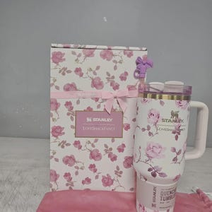 May include: A white Stanley Quencher tumbler with a floral rose pattern, a matching gift box, and a pink drawstring bag. The tumbler has a handle and a lid. The gift box is decorated with pink roses and a pink ribbon. The text "Stanley Loveshackfancy" is visible.
