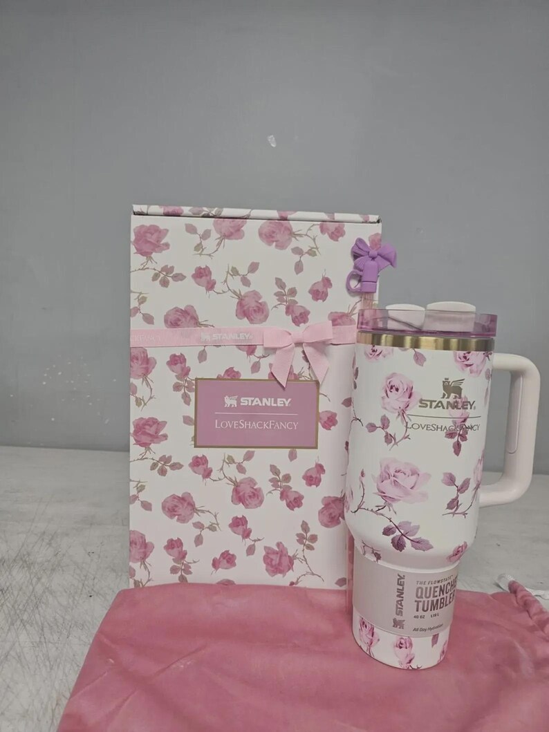 May include: A white Stanley Quencher tumbler with a floral rose pattern, a matching gift box, and a pink drawstring bag. The tumbler has a handle and a lid. The gift box is decorated with pink roses and a pink ribbon. The text "Stanley Loveshackfancy" is visible.