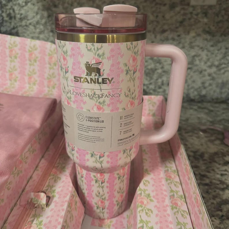 May include: A pink and floral Stanley Loveshackfancy travel mug with a handle. The mug has a pink lid and a floral pattern with pink roses. The mug is in its original packaging, which also has a matching floral pattern.