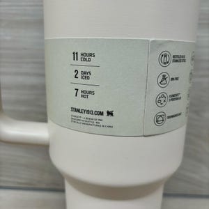 Pu&ograve; includere: Un bicchiere isolante bianco con manico. Il bicchiere ha un'etichetta con il testo "11 ORE FREDDO", "2 GIORNI CON GHIACCIO" e "7 ORE CALDO". L'etichetta include anche il testo "STANLEY1913.COM". Il bicchiere &egrave; realizzato in acciaio inossidabile riciclato e senza BPA.
