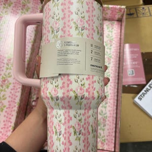 May include: A floral-patterned tumbler with a pink handle and lid. The tumbler features a white background with pink roses and green leaves. The lid has a gold rim. The text on the label reads "FLOWSTATE 3-POSITION LID".