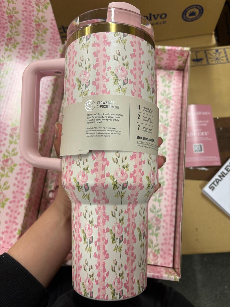 May include: A floral-patterned tumbler with a pink handle and lid. The tumbler features a white background with pink roses and green leaves. The lid has a gold rim. The text on the label reads "FLOWSTATE 3-POSITION LID".