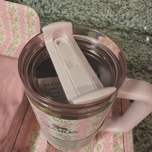 May include: A pink Stanley travel mug with a floral design. The mug has a white lid and handle. The mug is sitting on a pink floral patterned surface.