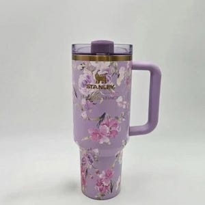 May include: A floral Stanley tumbler in lavender with a matching handle and lid. The tumbler features a gold rim and the Stanley logo. The floral pattern includes pink and purple flowers.