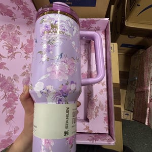 May include: A lavender Stanley Quencher tumbler with a floral design. The tumbler has a handle and a lid with gold accents. The floral pattern includes pink and purple flowers. The tumbler is in its original box, which has a matching floral pattern.