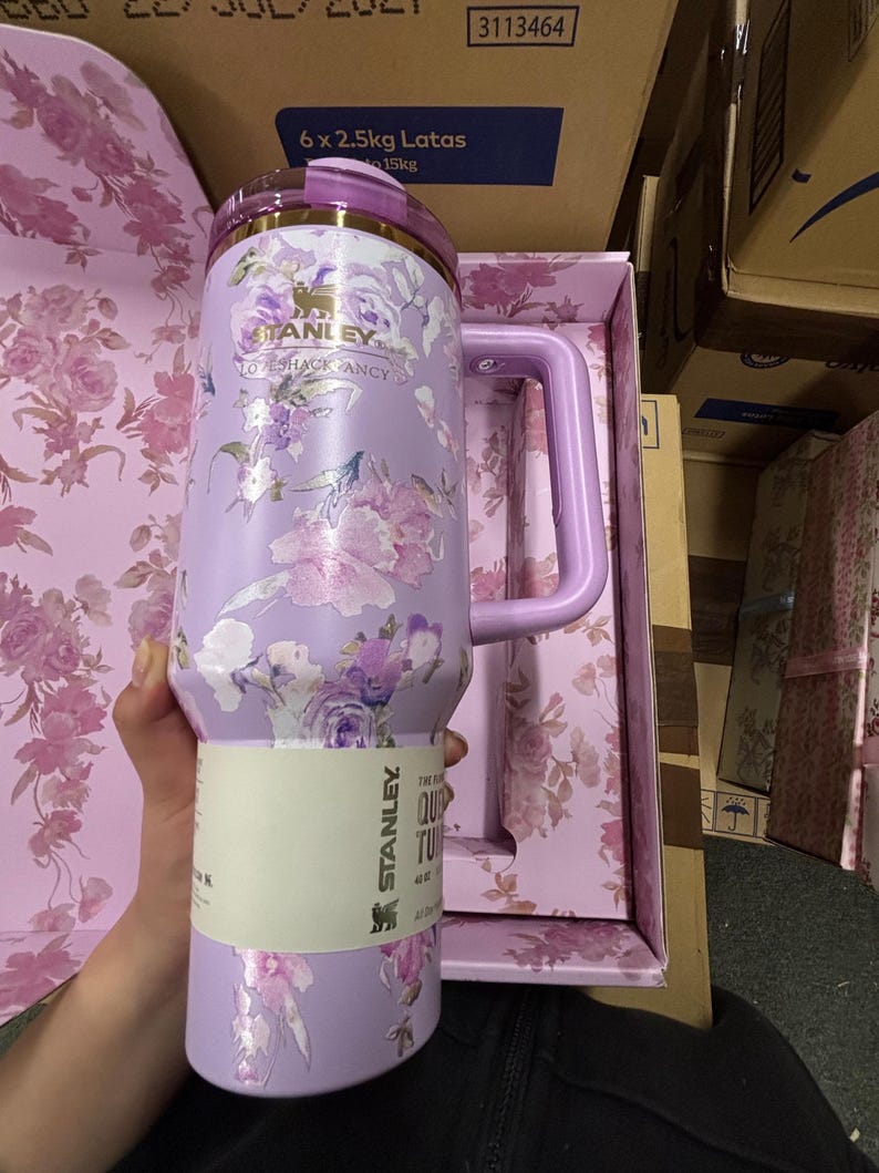 May include: A lavender Stanley Quencher tumbler with a floral design. The tumbler has a handle and a lid with gold accents. The floral pattern includes pink and purple flowers. The tumbler is in its original box, which has a matching floral pattern.