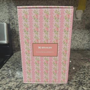May include: A pink box with a floral pattern of pink roses and green leaves. The box has the "STANLEY" and "LoveShackFancy" logos in a gold rectangle. The box is on a gray countertop.