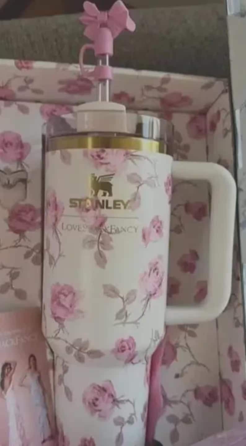 May include: White Stanley mug with a floral rose pattern and a gold rim. The mug has a white handle and a pink bow on the straw. The mug has the text "Stanley Loves Fancy".