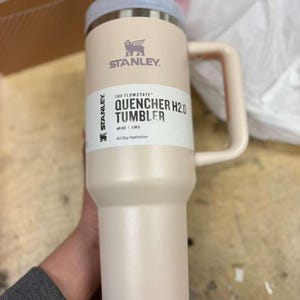 Pu&ograve; includere: Un bicchiere Stanley Quencher H2.0 beige con coperchio e manico azzurro. Il bicchiere presenta il logo Stanley e il testo "QUENCHER H2.0 TUMBLER". Contiene 1,18 L ed &egrave; progettato per l'idratazione quotidiana.