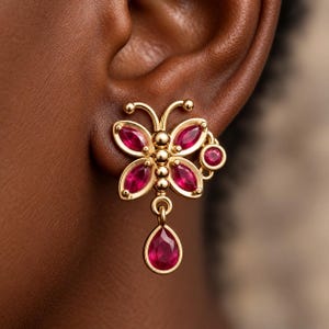 Puede incluir: Un pendiente dorado con forma de mariposa que presenta cuatro piedras rojas de talla marquesa formando las alas, una fila central vertical de pequeñas esferas doradas y una piedra roja en forma de pera que cuelga, con un pequeño acento redondo rojo.