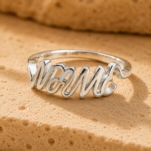 Könnte beinhalten: Ein silberfarbener Metallring mit dem Wort Mama in einer durchgehenden, fließenden Schreibschrift. Der Ring hat eine polierte, reflektierende Oberfläche und eine dünne, kreisförmige Ringschiene, die vorne in den Schriftzug übergeht.
