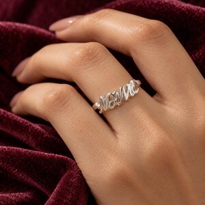Peut inclure: Une bague de couleur argentée portant le mot MAMA écrit dans une police cursive et fluide. Le métal présente une finition polie et réfléchissante qui capte la lumière, et l'anneau est fin et simple, conçu pour reposer sur un doigt.