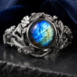 Può includere: Un bracciale rigido in argento con una pietra preziosa di labradorite rotonda centrale dai riflessi blu e gialli. La fascia presenta un design in rilievo dettagliato di rami d'albero strutturati, foglie di quercia e piccole ghiande attorno.