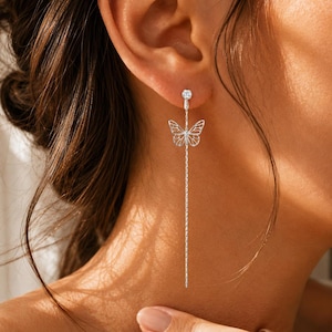 May include: A silver-toned drop earring featuring a small clear stud at the top, a delicate openwork butterfly charm suspended in the centre, and a thin vertical chain extending downwards from the butterfly to create a long, dangling silhouette.