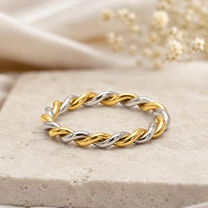 May include: A thin ring featuring a twisted design with alternating polished gold-tone and silver-tone metal strands. The band has a continuous rope-like texture created by the intertwined metallic wires, forming a circular shape with a smooth finish.