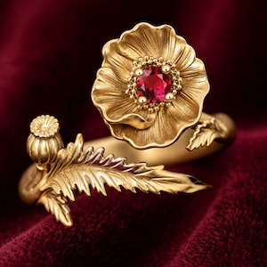 Op de afbeelding: Een gouden ring met een gebeeldhouwde bloem met een centrale ronde rode edelsteen omringd door kleine gouden kraaltjes. De ringband is gevormd als een stengel met een getextureerd blad en een kleine, afgeronde zaaddoos aan het uiteinde.