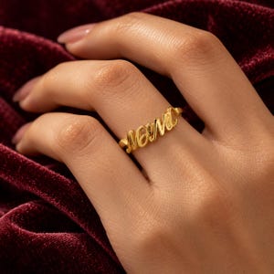 Può includere: Un anello color oro che presenta la parola MOM in un carattere corsivo e fluido. Le lettere sono collegate in un'unica linea continua, formando il pezzo centrale della fascia. Il metallo ha una finitura lucida e riflettente che cattura la luce.