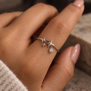 May include: A silver-coloured ring featuring a thin band adorned with two small four-pointed star shapes and a larger star shape. A single round, clear, faceted stone hangs from the largest star, creating a delicate dangling effect on the finger.