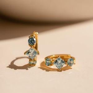 Può includere: Orecchini a cerchio color oro con pietre preziose azzurre. Un orecchino presenta una gemma rotonda in alto e una gemma quadrata in basso. L'altro ha tre gemme: due rotonde e una quadrata.