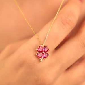 May include: A gold chain necklace featuring a four-leaf clover pendant. The clover is composed of four heart-shaped, vibrant pink gemstones set in gold, with a small gold stem extending from the bottom of the clover shape at the base of the pendant.