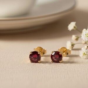May include: Two gold stud earrings, each featuring a round, deep red gemstone. The earrings are displayed on a neutral surface, with a blurred plate and small white flowers in the background. A classic jewellery item.