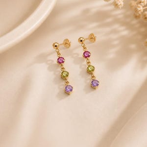 May include: A pair of gold drop earrings featuring three round faceted gemstones each. The stones are arranged vertically in a chain, displaying a vibrant color gradient of pink, light green, and purple. The earrings have a simple post back closure.
