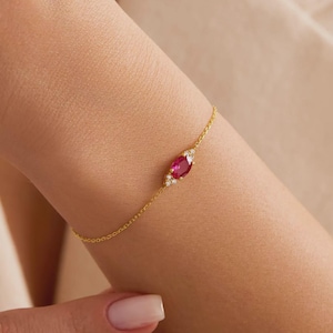 Könnte beinhalten: Ein zartes Armband aus einer Goldkette mit einem zentralen, marquise-geschliffenen, leuchtend pinken Edelstein, der auf beiden Seiten von zwei kleinen, klaren weißen Steinen flankiert wird. Die feinen Metallglieder bilden ein schmales Band.
