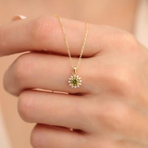May include: A gold chain necklace featuring a pendant with a central round olive-green gemstone surrounded by a halo of small, clear white stones arranged in a flower-like pattern. The delicate gold chain connects to the top of the floral pendant.
