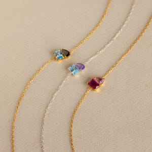May include: Three delicate necklaces with gemstone pendants. One features a gold chain with a blue and black stone, another a silver chain with blue and purple stones, and a third with a gold chain and pink and red stones.