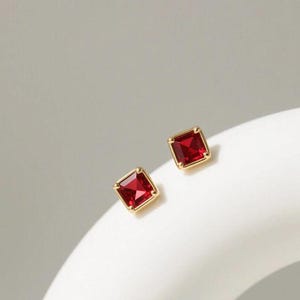 May include: A pair of square-cut red gemstone stud earrings set in gold metal. Each earring features a faceted deep red stone secured by four small prongs within a polished gold frame, creating a classic and symmetrical jewelry design.