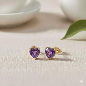 May include: A pair of heart-shaped stud earrings. Each earring features a vibrant purple gemstone set in a gold-colored metal. The earrings are displayed on a neutral-toned surface, with soft lighting.