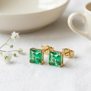 May include: A pair of stud earrings featuring square-cut, vibrant green gemstones set in gold-toned metal. Each earring has a four-prong setting and a standard post back, showcasing a clear, faceted surface that reflects light across the stone.