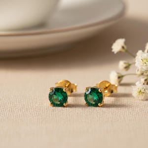 May include: A pair of gold stud earrings with round, emerald-green gemstones. The earrings are set in a four-prong setting. The background is a neutral beige color with a white flower and a white teacup.
