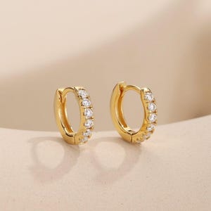Könnte beinhalten: Ein Paar goldene Creolen mit einer Reihe kleiner, klarer, rund geschliffener Steine, die entlang der Vorderkante jedes kreisförmigen Rings eingelassen sind. Die Ohrringe haben eine polierte Metalloberfläche und einen Klappverschluss.