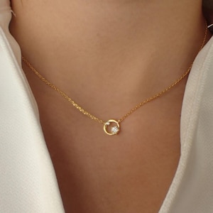 May include: A delicate gold necklace featuring a small, open circle pendant with a single, sparkling gemstone. The necklace has a fine chain and is worn around the neck, against a white shirt.