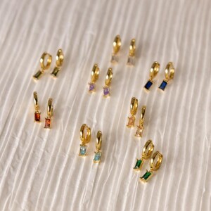Könnte beinhalten: Eine Sammlung goldener Creolen, die jeweils mit einem kleinen, rechteckigen Baguette-geschliffenen Edelsteinanhänger versehen sind. Das Set enthält Paare in verschiedenen Farben, darunter Rot, Orange, Hellblau, Grün, Dunkelblau, Violett und Transparent.