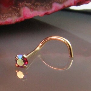 May include: A gold-colored nose ring with a curved design. The nose ring features a small, square-cut, iridescent gemstone that reflects multiple colors. The jewelry is set against a reflective surface.