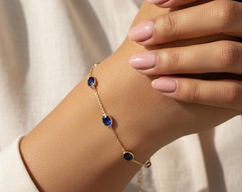 May include: A delicate gold bracelet featuring oval-shaped, deep blue gemstones. The bracelet is worn on a wrist, showcasing the gemstones' vibrant color against the gold chain. The bracelet is a piece of jewelry.
