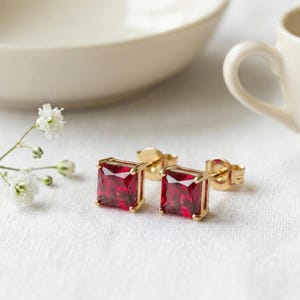 May include: A pair of stud earrings featuring square-cut, deep red gemstones set in polished gold-toned metal. Each earring has a secure post back mechanism, showcasing a faceted surface that reflects light, presented with small white flowers.